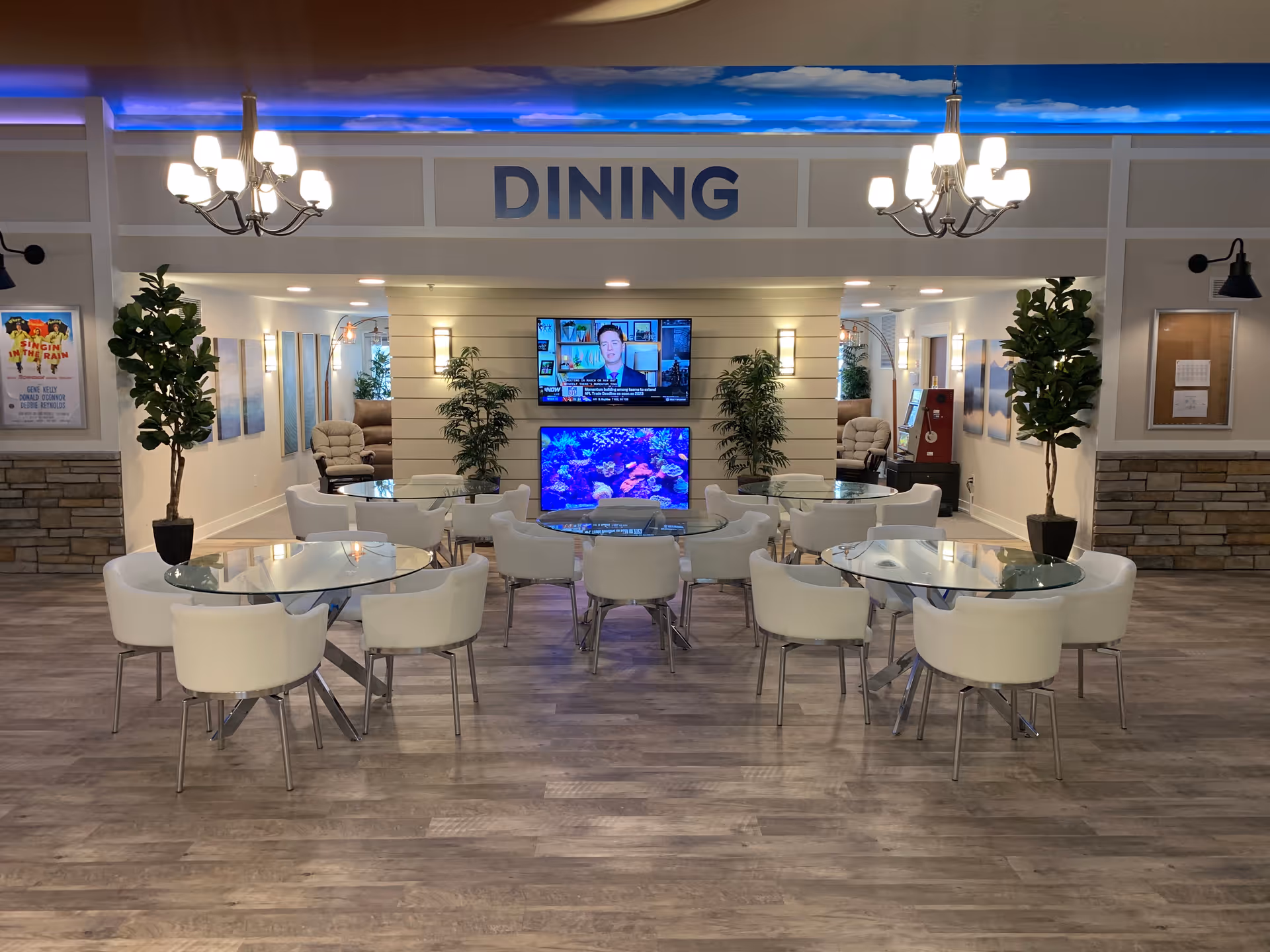 A dining area with several round glass tables surrounded by white chairs. The word 'DINING' is displayed prominently above the entrance. The room features wood flooring, potted plants, two chandeliers, and a wall with two televisions, one showing a news anchor and the other displaying an aquarium scene. There are also comfortable chairs and framed pictures along the walls.