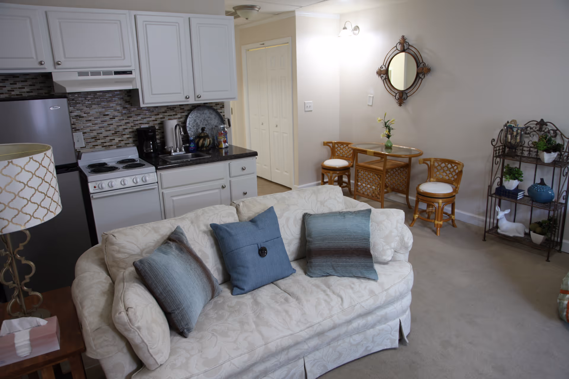 A cozy living area with a white patterned sofa adorned with three blue and gray cushions. Behind the sofa is a compact kitchen with white cabinets, a small stove, a refrigerator, and a black countertop with a coffee maker and other kitchen items. To the right, there is a small dining area with a round glass-top table and two wicker chairs with white cushions. A decorative mirror hangs on the wall above the table, and a metal shelf unit with plants and decorative items is placed against the wall. The room is softly lit by a wall-mounted light fixture.