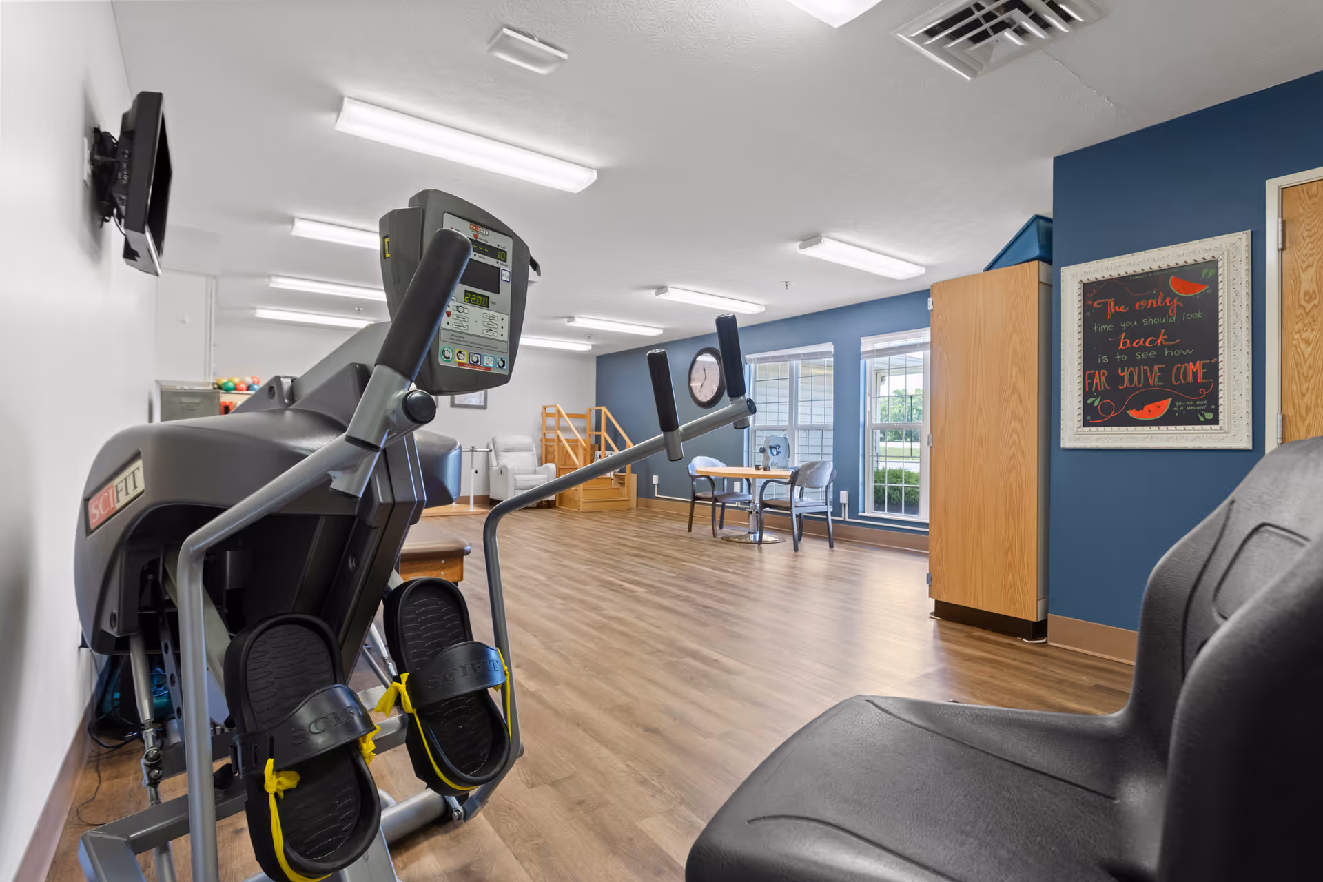 A spacious exercise room with wooden flooring and blue accent walls. The room contains exercise equipment including a SciFit machine in the foreground, a small table with two chairs near large windows, and a wooden staircase for physical therapy. A framed motivational quote is hung on the wall, and a wall-mounted TV is visible on the left side.