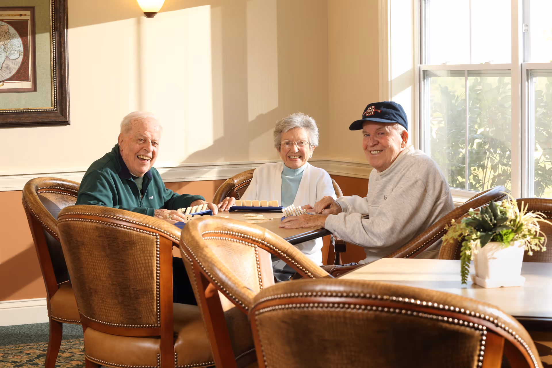 Three elderly people sitting around a table in a well-lit room playing a game and smiling. The room has large windows, framed artwork on the wall, and comfortable leather chairs. A small potted plant is on a nearby table.