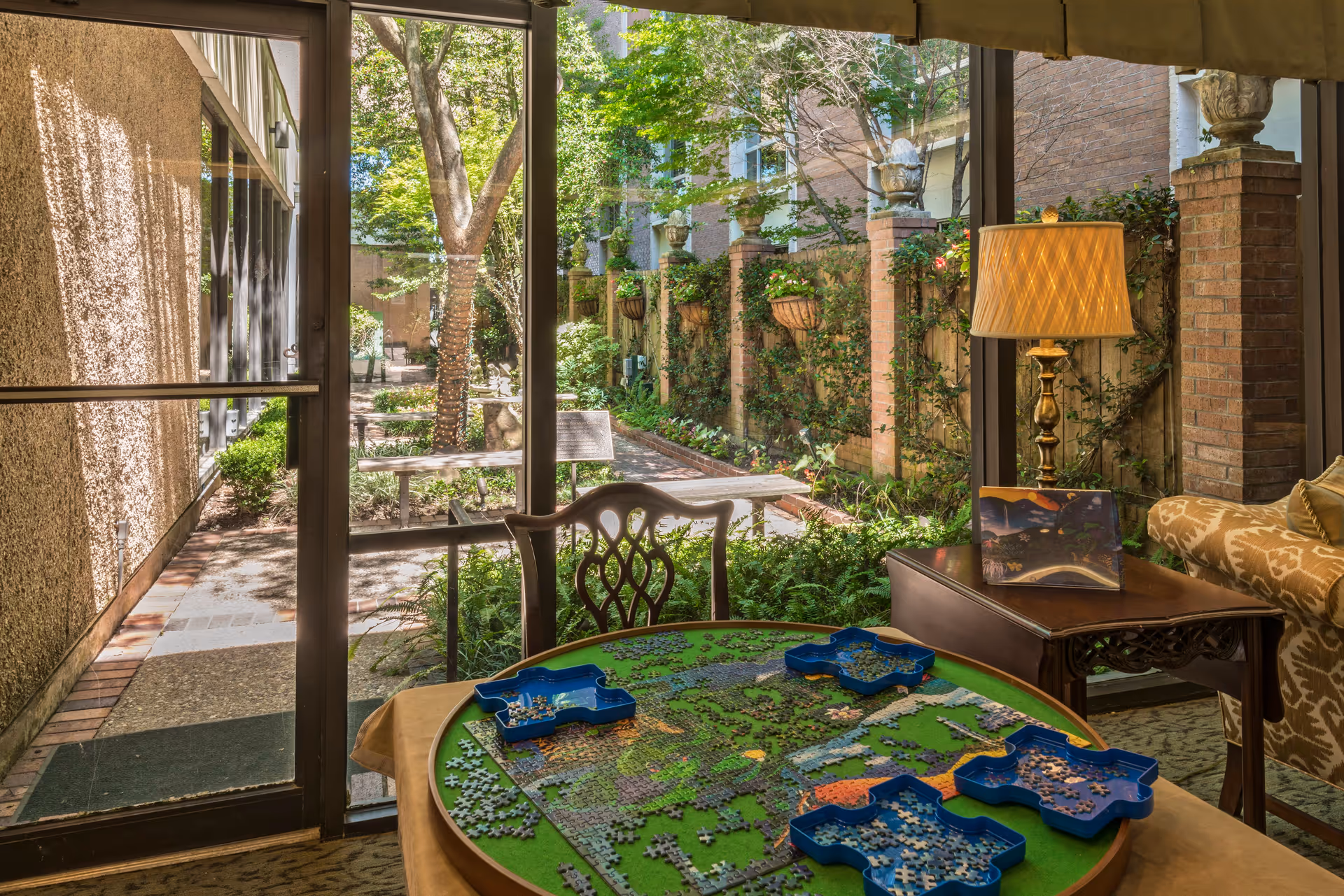 A cozy indoor seating area with a round table featuring a partially completed jigsaw puzzle and blue puzzle piece trays. Behind the table is a wooden chair and a side table with a lit lamp and a decorative item. Large glass doors open to a sunny outdoor garden with trees, plants, and a brick wall with hanging planters.