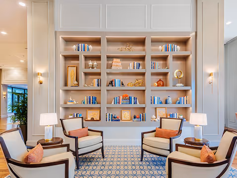 A bright and elegant sitting area with four cushioned armchairs arranged around two small round tables, each table holding a lamp. Behind the chairs is a large built-in bookshelf filled with decorative items and books. The room features soft lighting, a patterned carpet, and light-colored walls with panel detailing.