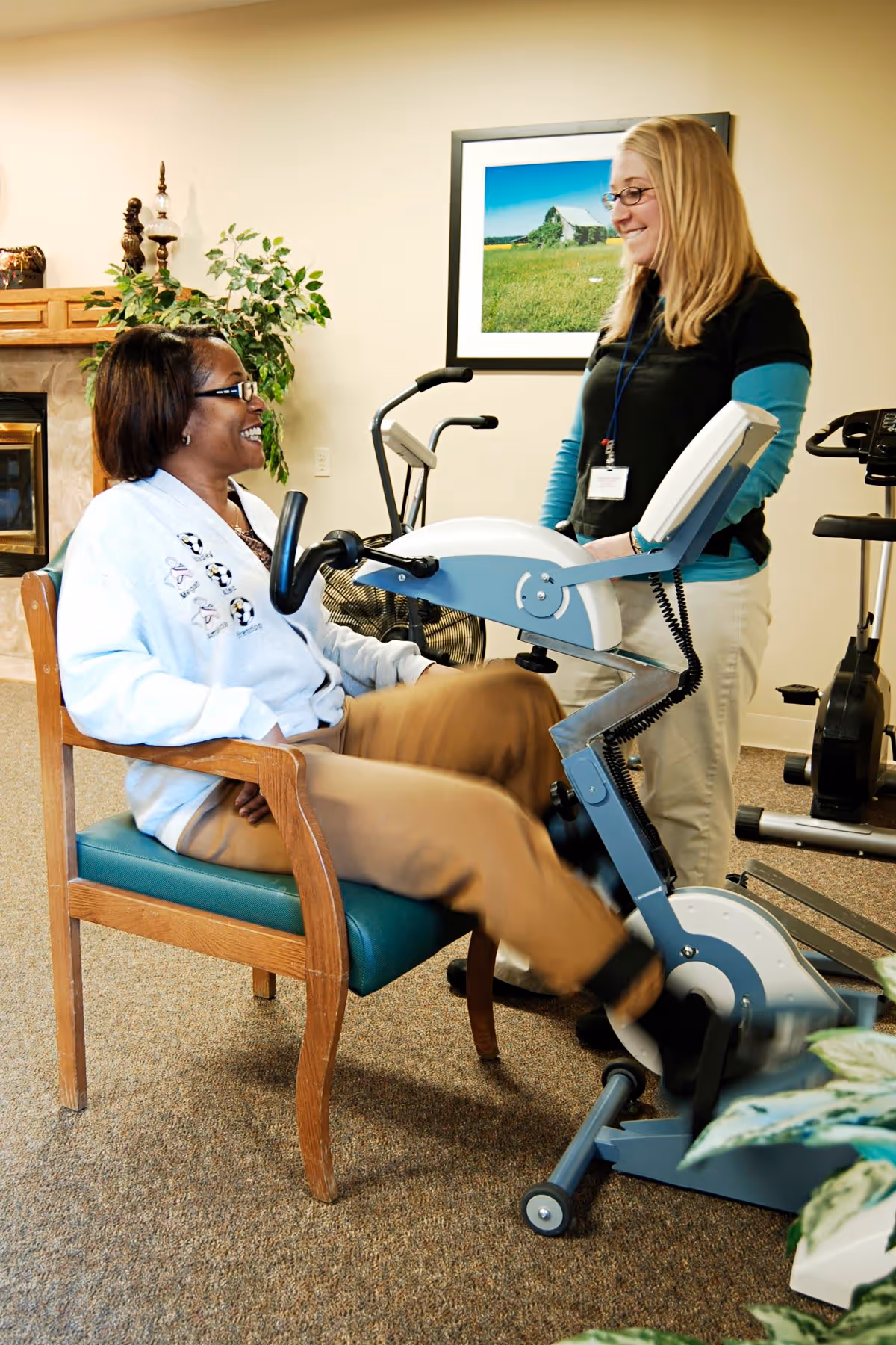 A woman seated on a chair is using a stationary exercise bike while another woman, standing nearby and wearing a name badge, smiles and looks on. The room has exercise equipment, a framed picture on the wall, a plant, and a fireplace in the background.