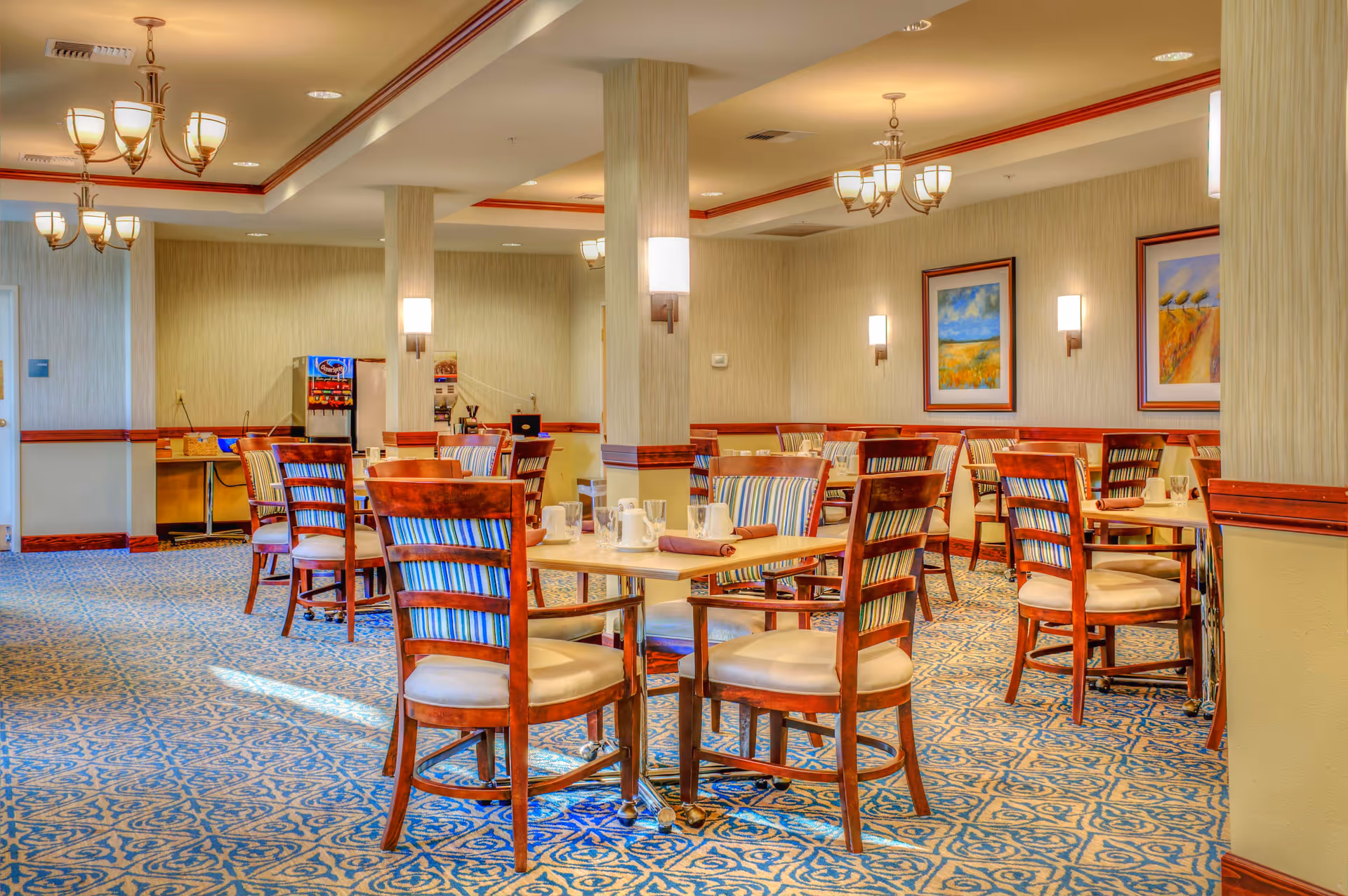 Bright communal dining room with wooden tables and striped upholstered chairs, chandeliers, and framed artwork on the walls.