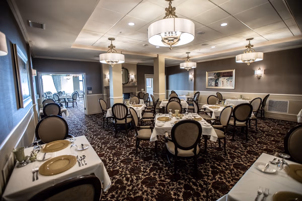 A formal dining room with multiple tables covered in white tablecloths, each set with plates, silverware, and glasses. The room features upholstered chairs, patterned carpet, elegant chandeliers, and wall sconces. There is a fireplace and a framed painting on the wall, with an adjoining room visible in the background containing more dining tables and chairs.