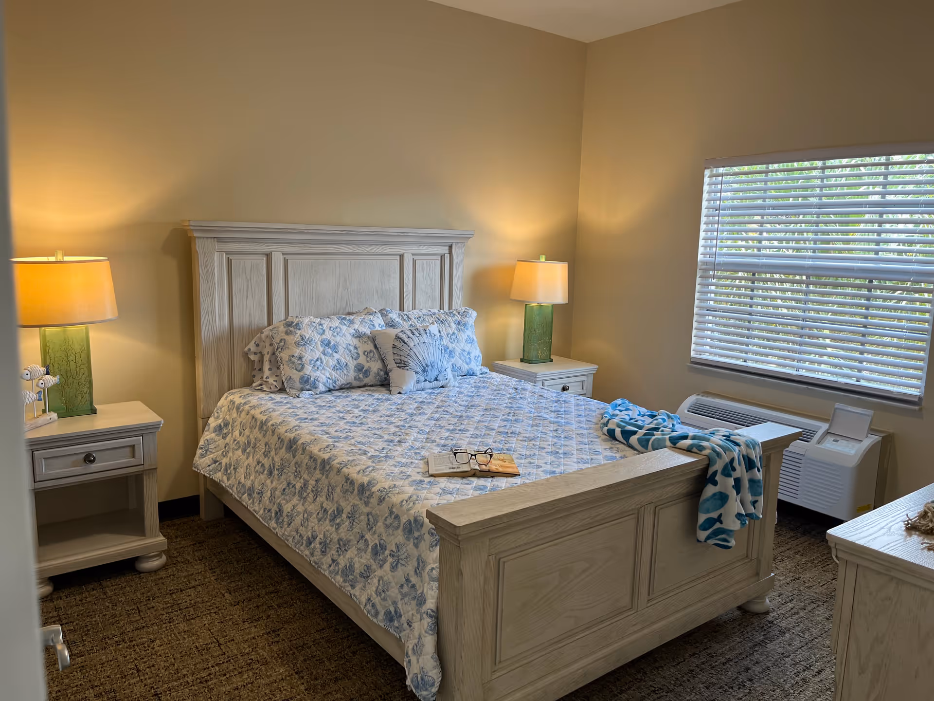 A cozy bedroom with a wooden bed frame and matching nightstands on either side. The bed is made with a blue and white patterned quilt and pillows. Two lamps with green bases and beige shades sit on the nightstands. A window with blinds is on the right wall, and a folded blue and white blanket is draped over the footboard of the bed. The room has beige walls and carpeted flooring.