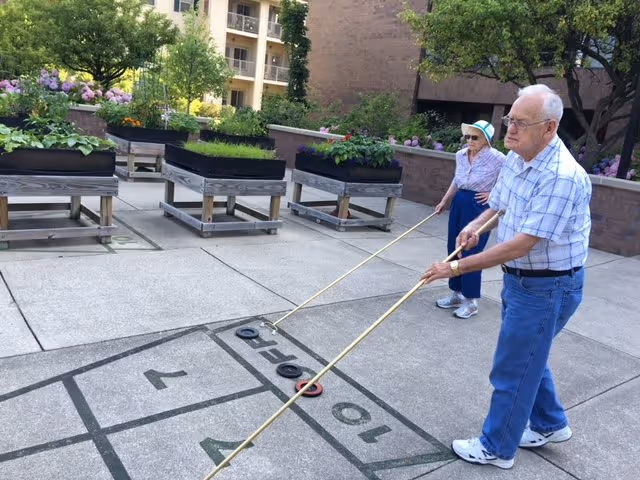 Two seniors playing shuffleboard on an outdoor courtyard court with planter boxes and building balconies in the background.