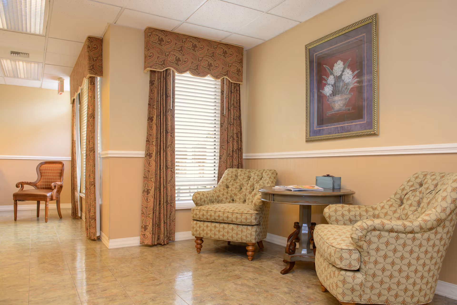 A seating area with two patterned armchairs and a round table beside a window with drapes and framed artwork on the wall.