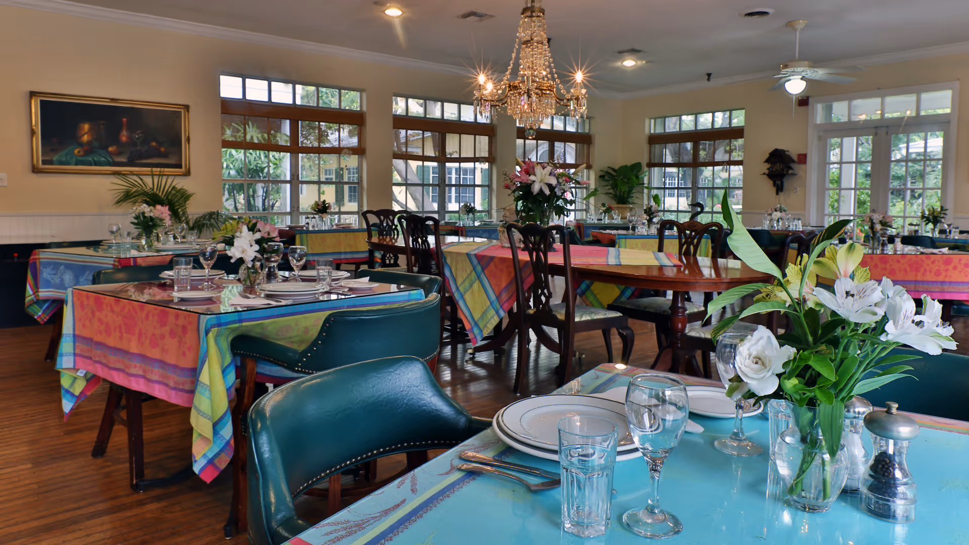 A bright and elegant dining room with multiple tables covered in colorful tablecloths. Each table is set with plates, glasses, silverware, and floral centerpieces. Large windows allow natural light to fill the room, and a crystal chandelier hangs from the ceiling. The room has wooden floors and a cozy, inviting atmosphere.