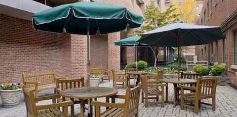Outdoor courtyard with wooden tables, chairs, and green umbrellas between brick buildings.