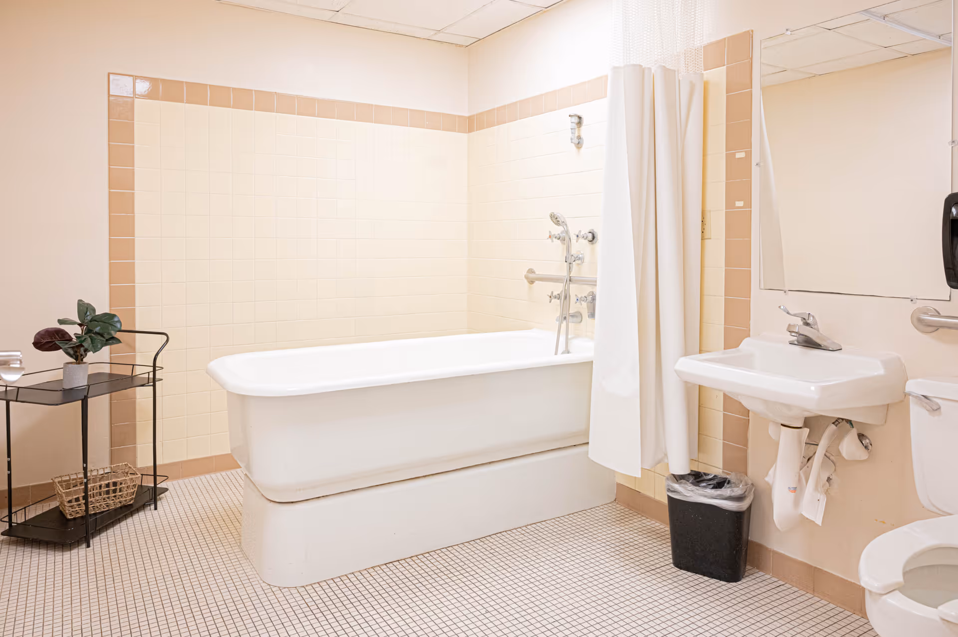 A clean bathroom with a white bathtub, a shower curtain, a white sink with a faucet, a mirror above the sink, a toilet, a small black trash bin, and a black metal shelf holding a potted plant and a basket. The walls are tiled in beige and cream colors, and the floor has small square tiles.