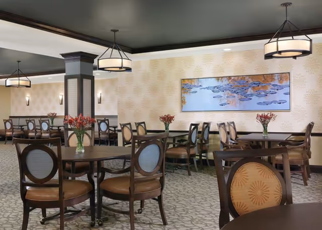 A dining room with multiple round and rectangular tables surrounded by wooden chairs with cushioned seats and backs. Each table has a vase with red flowers. The room features patterned wallpaper, carpeted floor, and modern hanging light fixtures. A large framed artwork depicting water lilies is mounted on the wall.