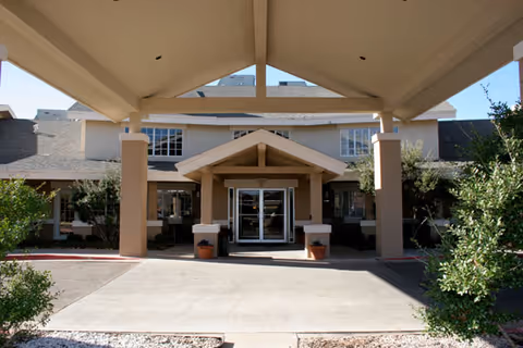 Front entrance of a senior living facility with a covered drop-off area supported by columns, two potted plants near the entrance, and a two-story building with multiple windows in the background.
