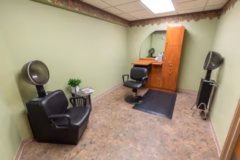 Small room with light green walls and a brown patterned border near the ceiling. The room contains two black salon chairs with hair dryers attached, a small table with a plant and magazines, and a wooden cabinet with a mirror and hair care products.