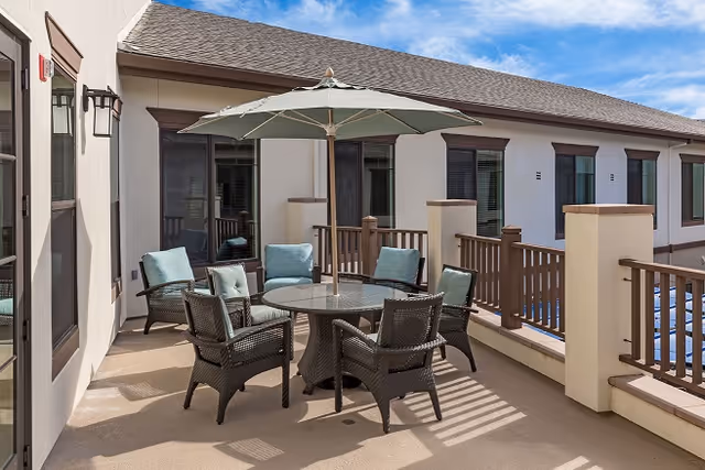 Outdoor patio area with a round glass table surrounded by six wicker chairs with cushions and a large umbrella providing shade. The patio is adjacent to a building with multiple windows and a railing along the edge.