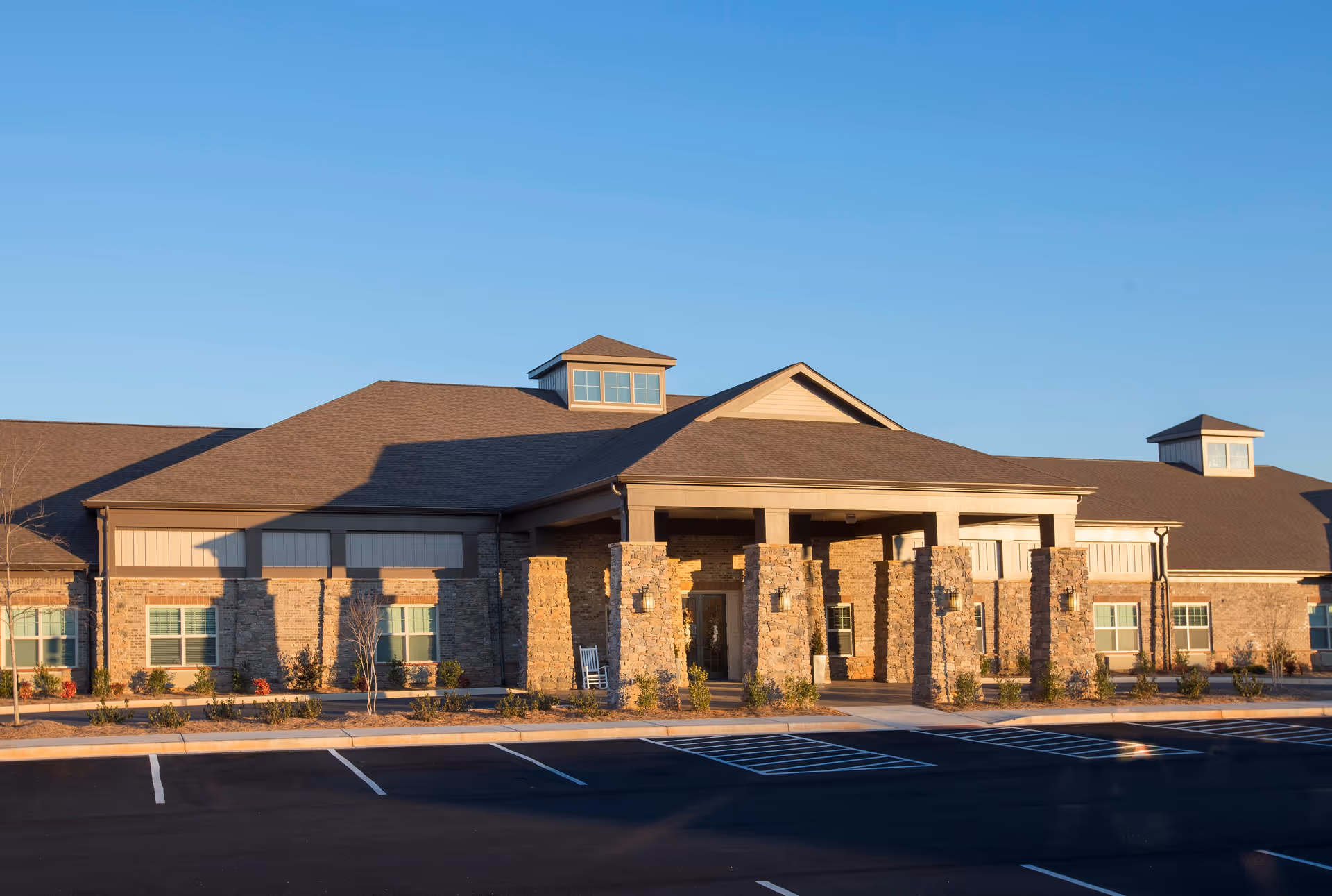 Front exterior of a single-story senior living facility with stone columns, a covered entrance, and an empty parking lot under a clear blue sky.
