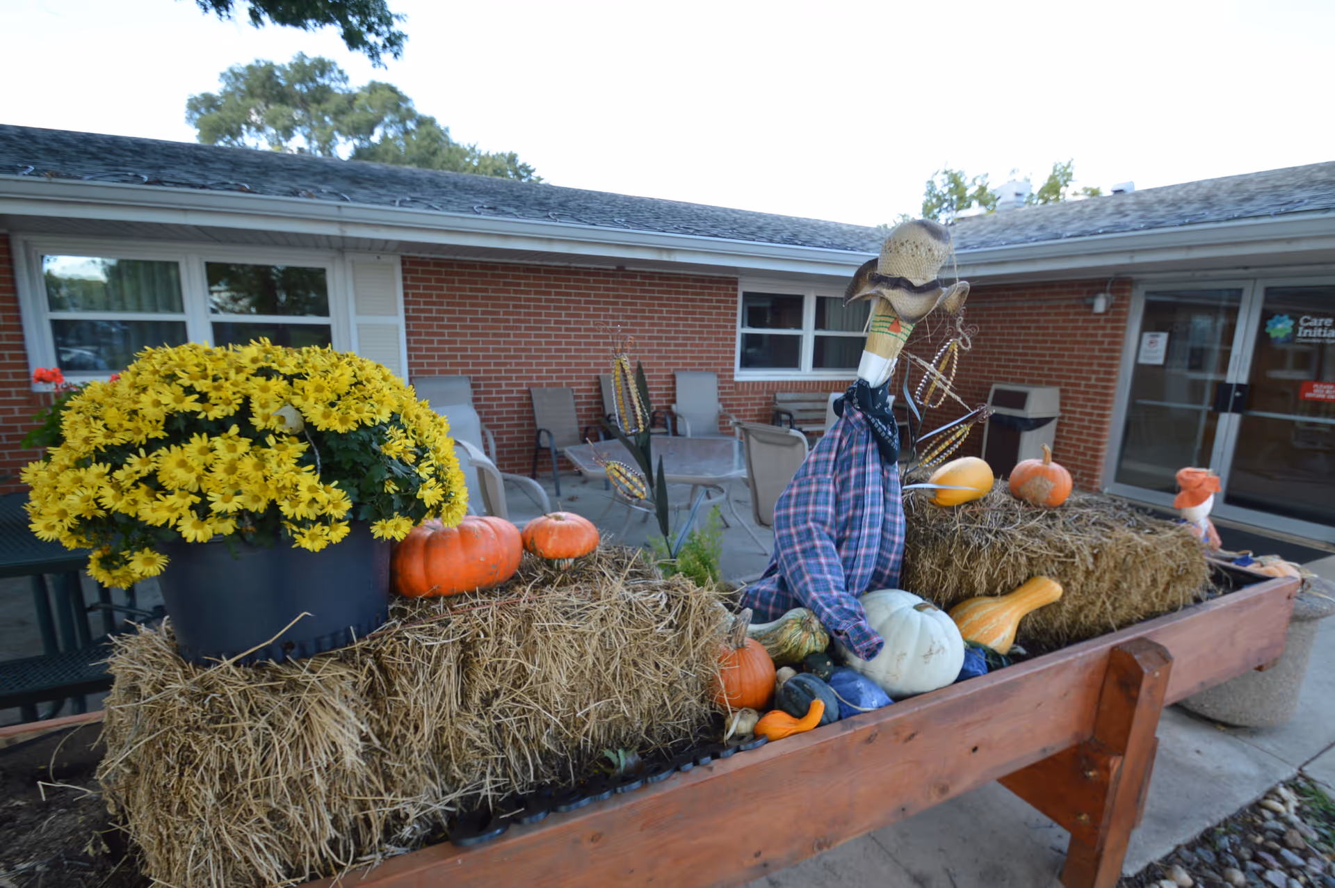 Outdoor autumn-themed decoration with hay bales, pumpkins, gourds, yellow flowers, and a scarecrow figure dressed in a plaid shirt and hat, set in front of a brick building with patio chairs and tables.