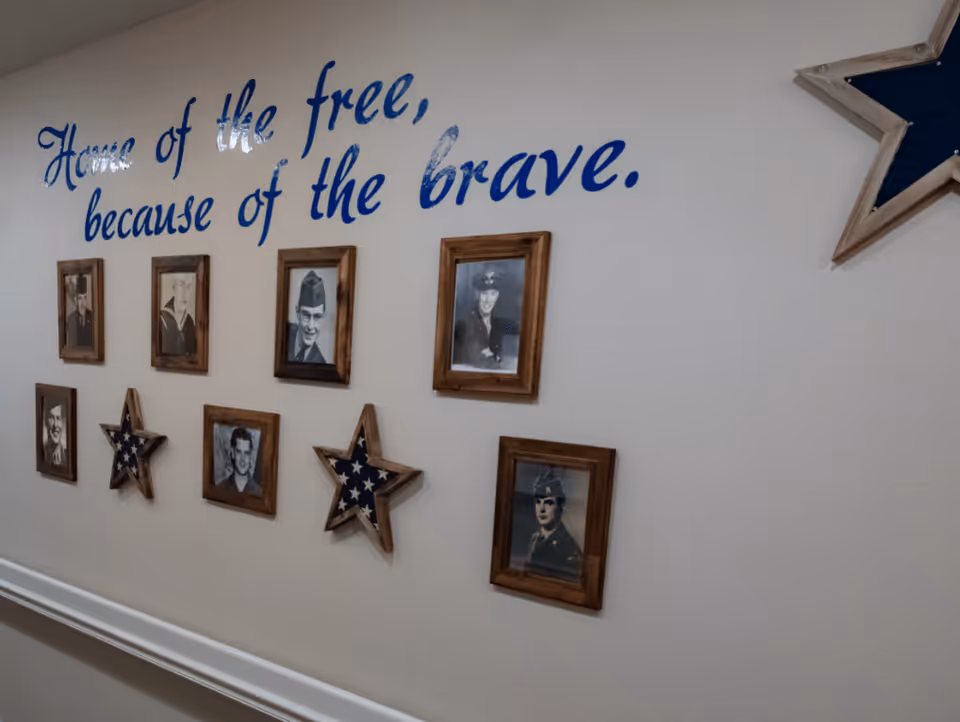 A wall display with the phrase 'Home of the free, because of the brave.' in blue cursive letters above framed black and white photographs of military personnel and wooden stars containing folded American flags.