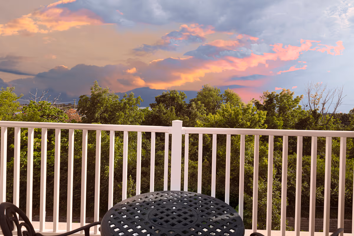 View from a balcony showing a white railing and patio table overlooking treetops under a colorful sunset sky.