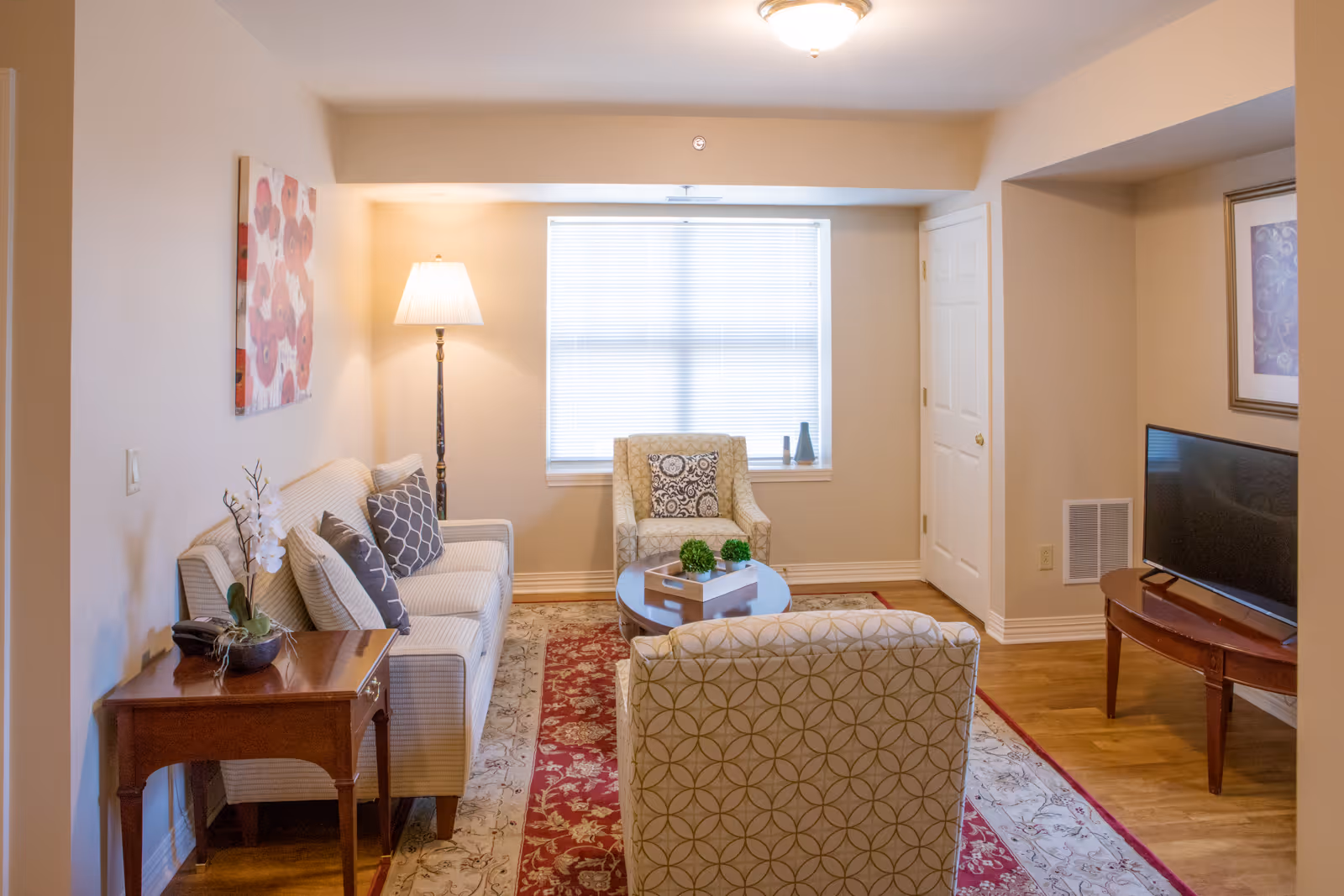 A cozy living room with a beige sofa, two patterned armchairs, a round coffee table with decorative plants, a floor lamp, a wooden side table with a potted orchid, a flat-screen TV on a wooden stand, and a window with blinds letting in natural light. The room has light-colored walls and a red and beige patterned area rug.