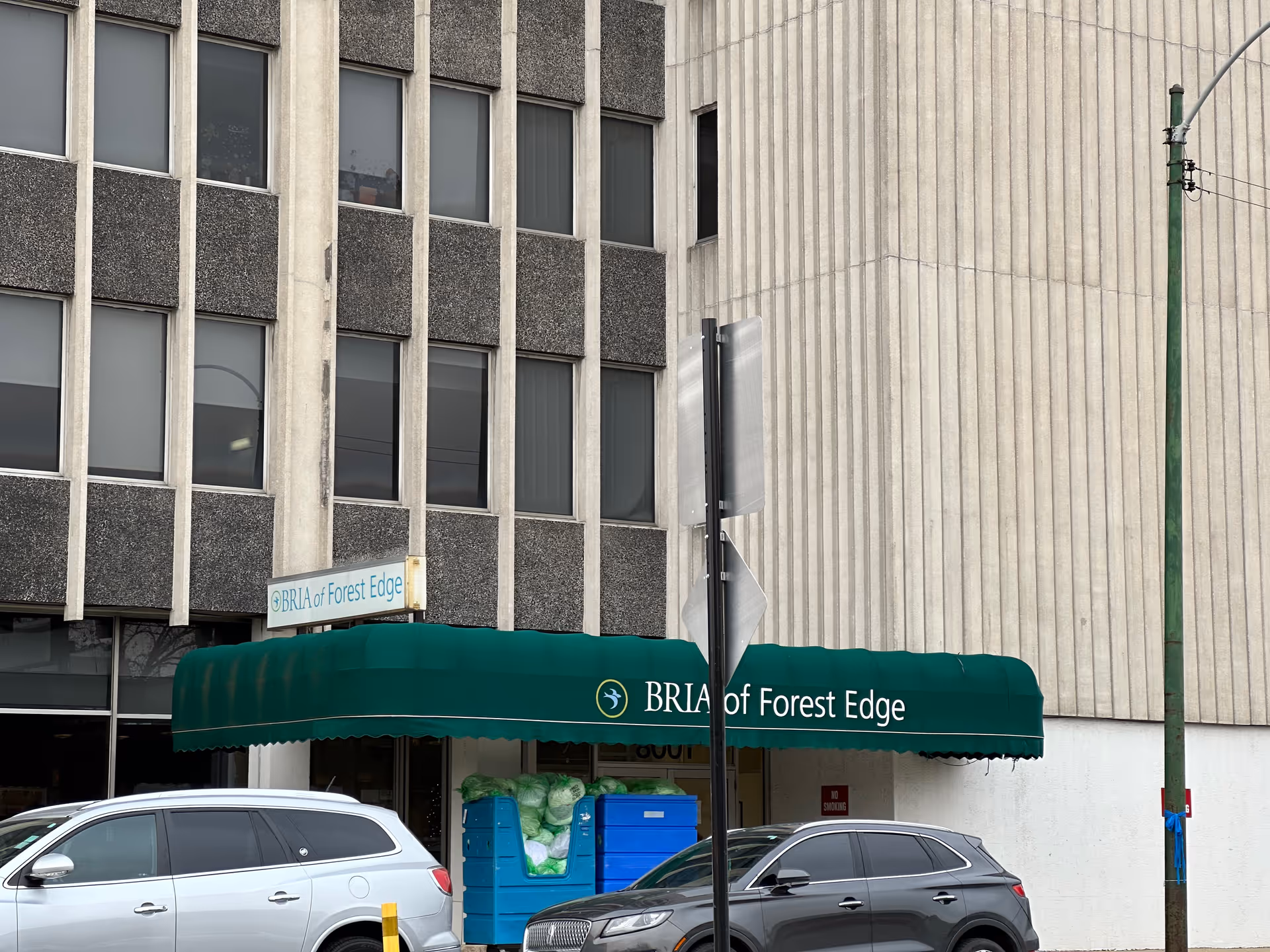 Exterior view of a multi-story building with a green awning that reads 'BRIA of Forest Edge'. Two cars are parked in front of the building, and there are some blue and green bins near the entrance.