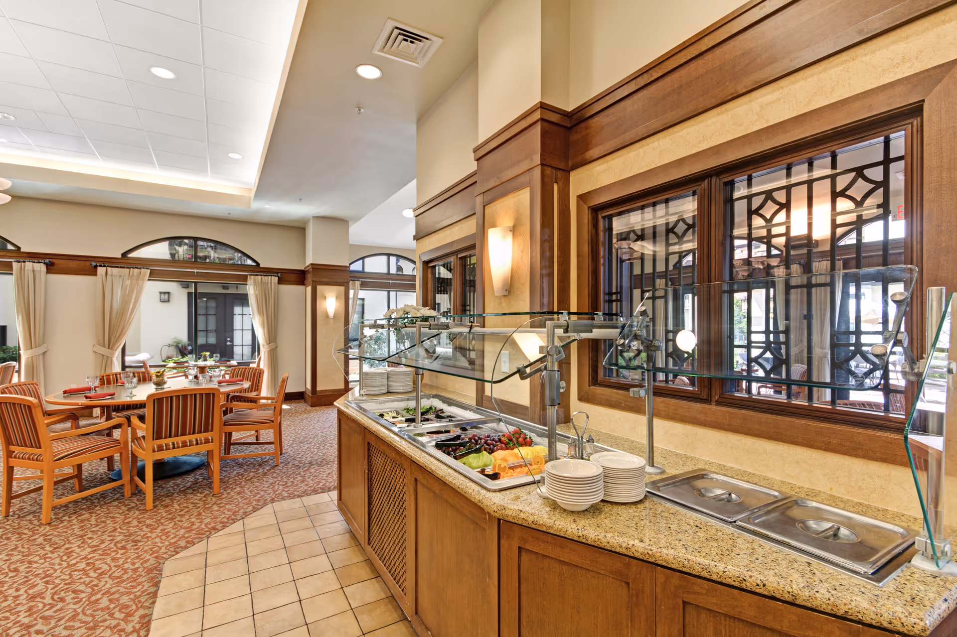 Interior view of a dining area in a senior living facility featuring a buffet counter with plates and a variety of fresh fruits and salad items. The room has wooden furniture, round tables with chairs, large windows with decorative grills, and beige walls with wood paneling and soft lighting.