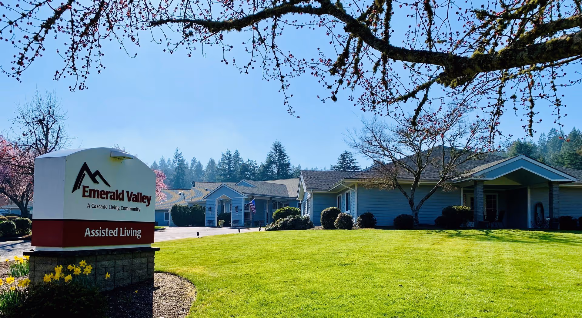 Exterior view of Emerald Valley Assisted Living facility with a large green lawn, trees with budding branches, and a clear blue sky. The building is a single-story structure with a covered entrance and several windows. A sign in the foreground reads 'Emerald Valley A Cascade Living Community Assisted Living.'