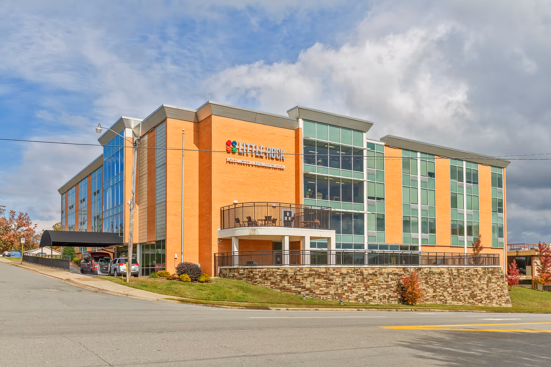 Three-story brick-and-glass senior care facility with a covered drop-off, terrace balcony, and parked cars under a partly cloudy sky.