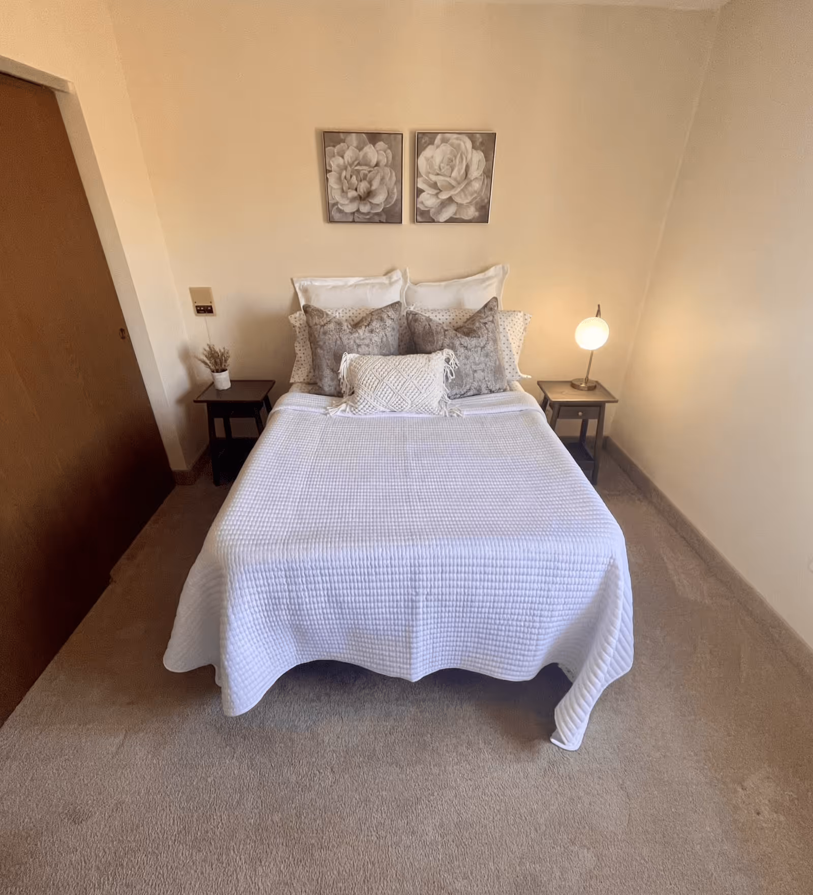 Neatly made bed with a white quilt, decorative pillows, two nightstands and a lamp in a small bedroom.