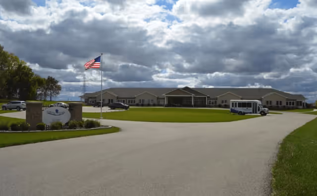 Wide view of Autumn Trace Attica Assisted Living facility showing a single-story building with a large driveway and green lawn in front. An American flag is flying on a flagpole near the entrance sign, and a small shuttle bus is parked on the driveway. The sky is partly cloudy.