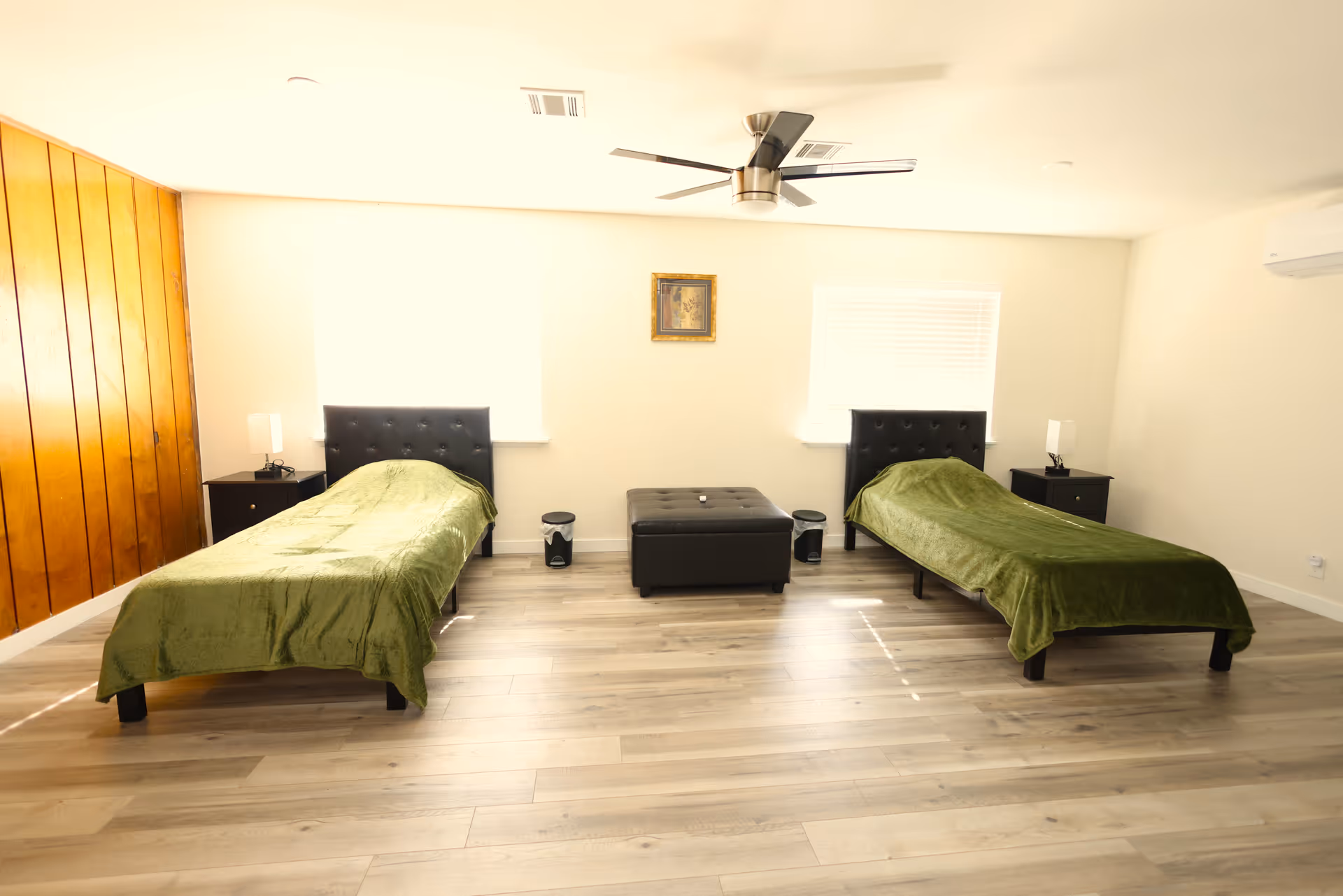 A bright bedroom with two single beds covered with green blankets, each bed has a black tufted headboard. Between the beds is a black ottoman and two small black nightstands with lamps. The room has light wood flooring, a ceiling fan, and two windows with white blinds. One wall is covered with wooden paneling.