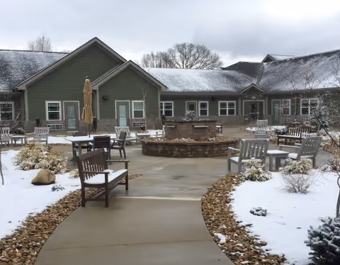 Outdoor courtyard area of a senior living facility with a circular stone fire pit in the center, surrounded by multiple wooden benches and chairs. The ground is partially covered with snow, and the building with green siding and multiple windows is visible in the background under a cloudy sky.