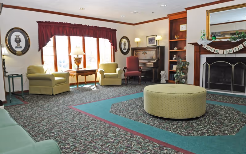A cozy living room with patterned carpet, a large round ottoman in the center, two green armchairs near a window with red curtains, a red upholstered chair, a wooden piano, built-in wooden shelves, and a fireplace with a mirror above it and a decorative banner.