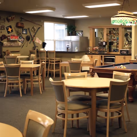 A cozy common area with multiple round and square tables surrounded by chairs, a pool table with balls set up, a kitchen counter with bar stools, a refrigerator, microwave, and cabinets. The walls are decorated with various vintage items and signs, and a stained glass hanging lamp is above the pool table.