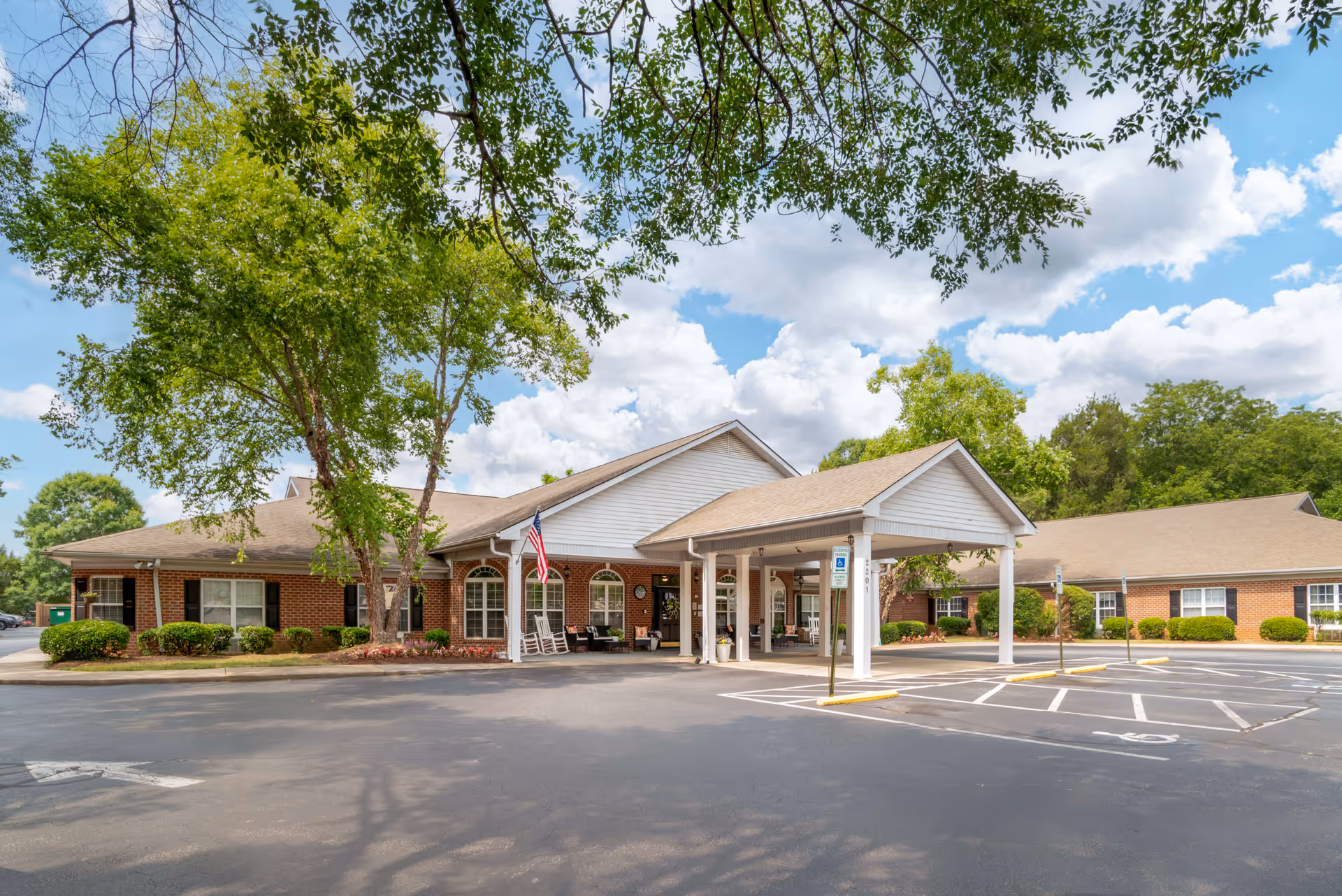 Exterior view of a single-story brick building with a covered entrance, surrounded by trees and greenery under a partly cloudy sky. There is a parking lot with marked spaces in front of the building.