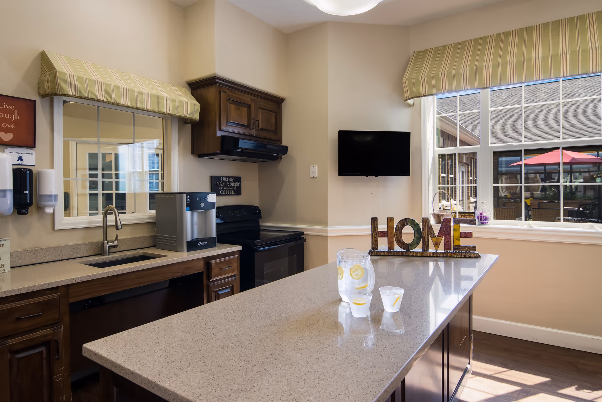 A cozy kitchen area with wooden cabinets, a countertop island with a pitcher and two glasses of lemon water, a small wall-mounted TV, and a window with striped valances showing an outdoor patio with red umbrellas.