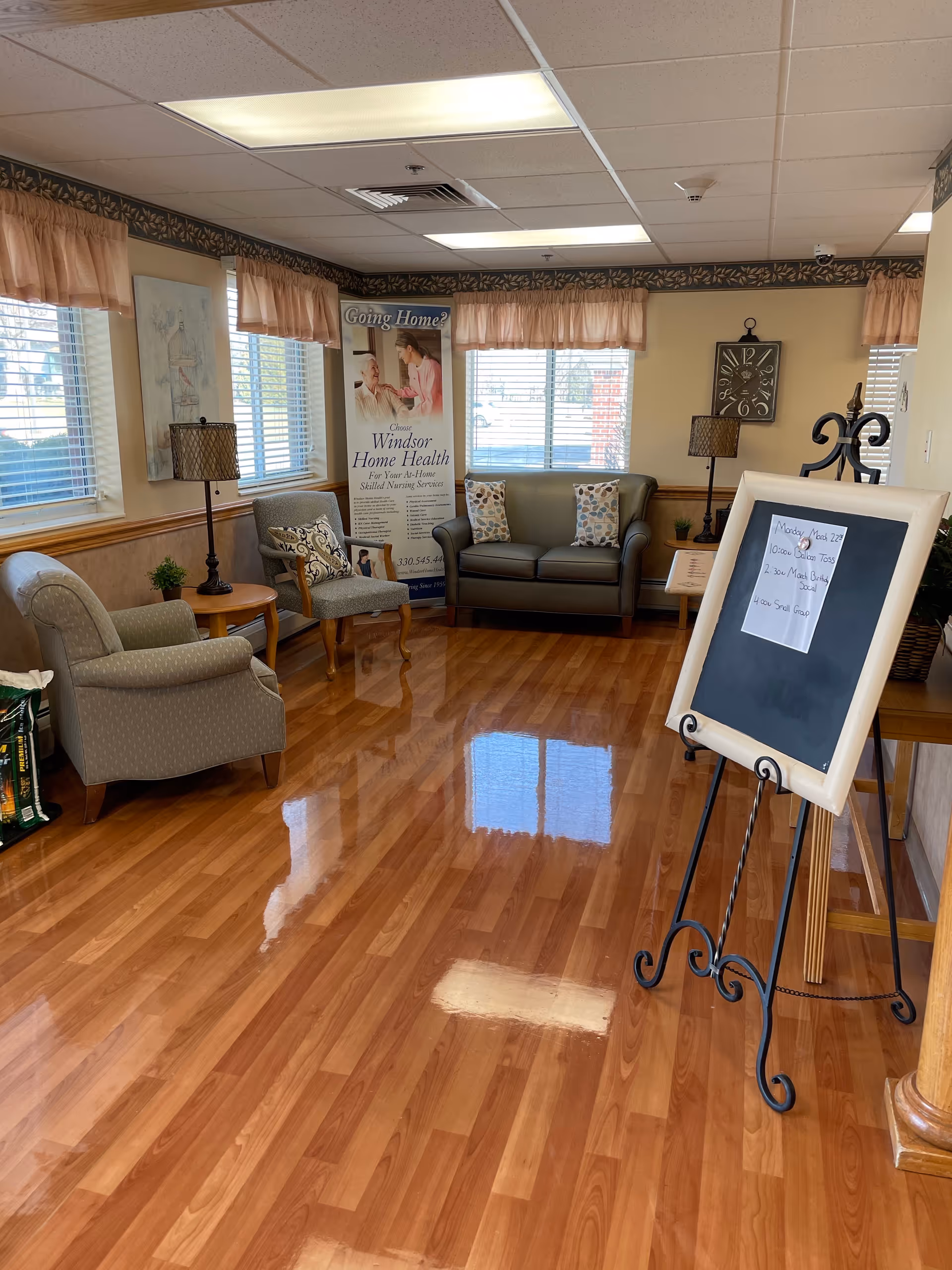 A cozy living room area with wooden flooring, a gray sofa with patterned pillows, two armchairs, a small round wooden table with a lamp, and a standing blackboard with a paper listing daily activities. The room has several windows with peach-colored valances, a wall clock, and a banner promoting Windsor Home Health skilled nursing services.