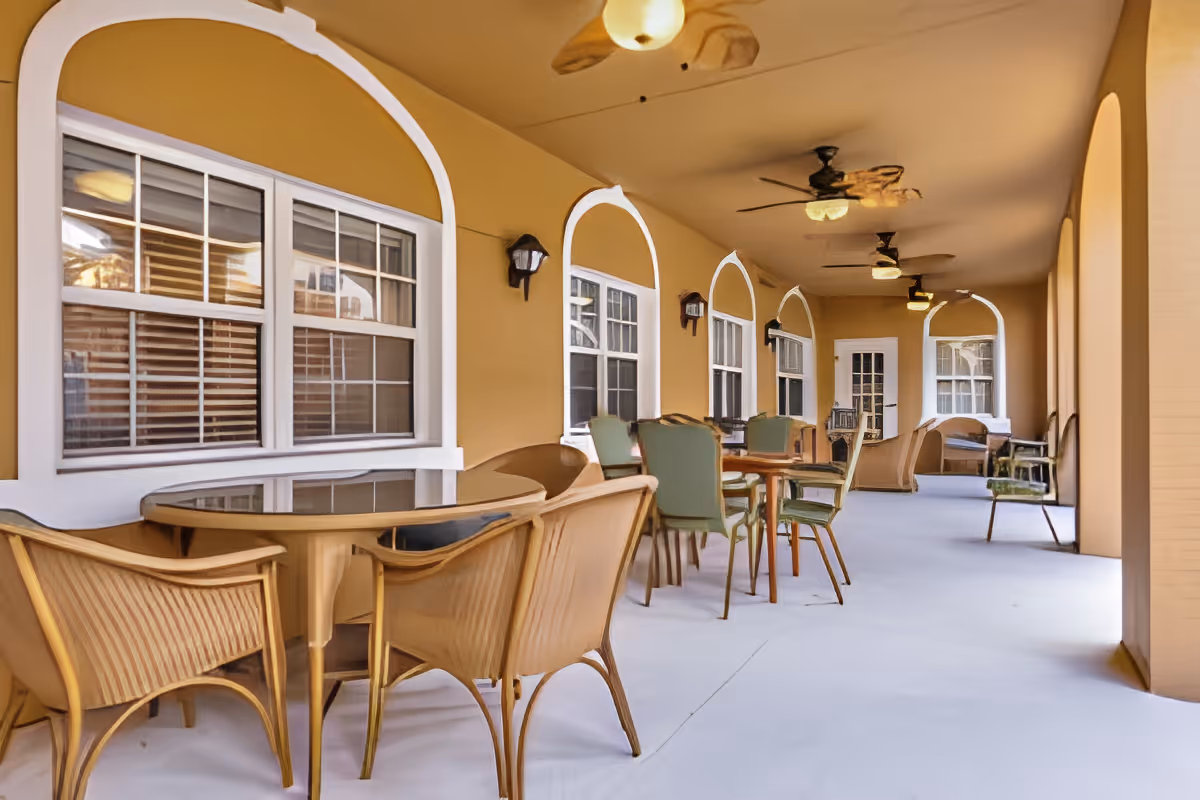 Covered outdoor patio area with multiple tables and chairs arranged for seating. The walls are painted a warm beige color with white trim around the windows and doors. Ceiling fans and light fixtures are mounted on the ceiling, providing ventilation and lighting. The space appears clean and inviting for socializing or relaxing.