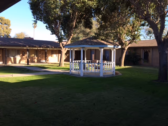 A sunny courtyard with a white gazebo surrounded by grass, trees, and single-story brick buildings.