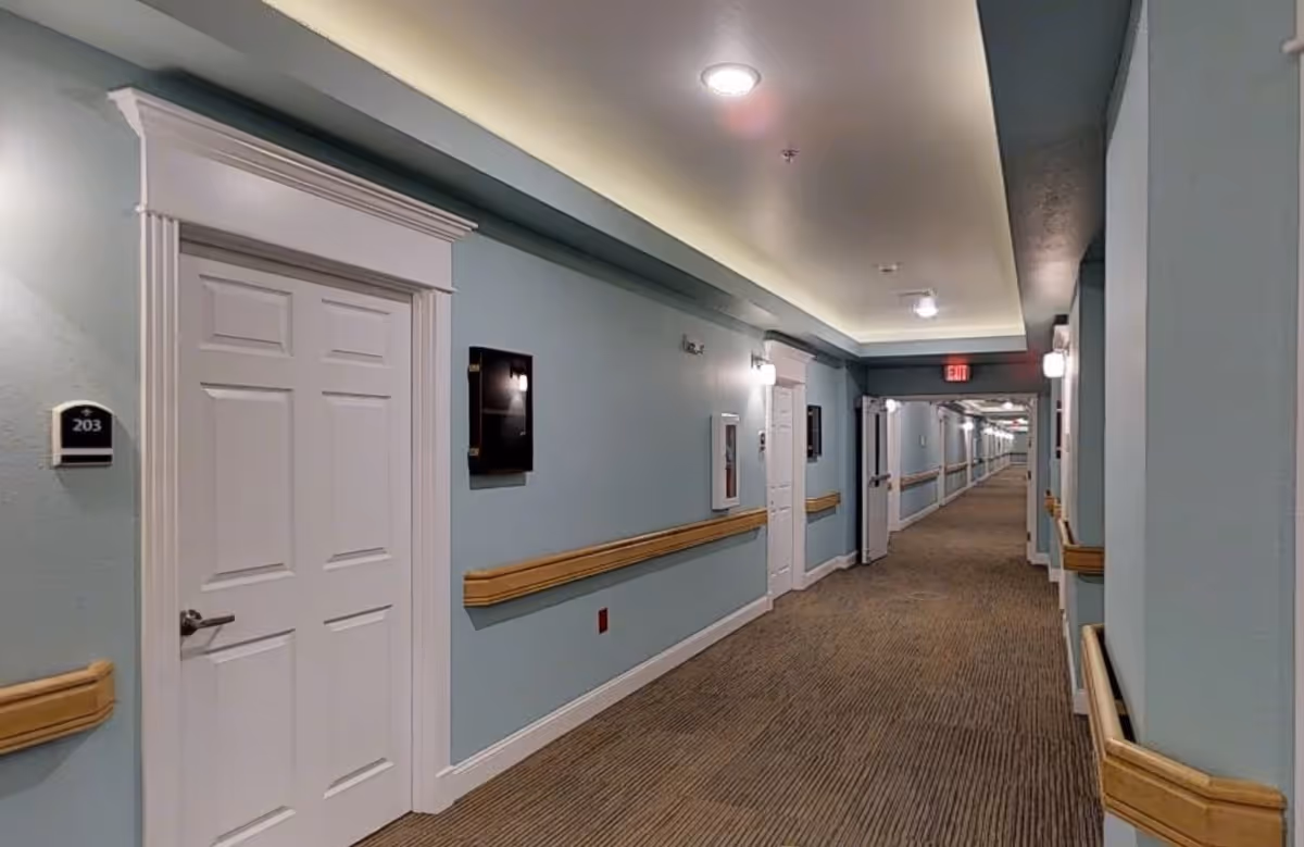 A long, well-lit hallway in a senior living facility with light blue walls, white doors, and wooden handrails on both sides. The carpet is brown with a subtle pattern, and there are wall-mounted lights and an exit sign visible at the far end.