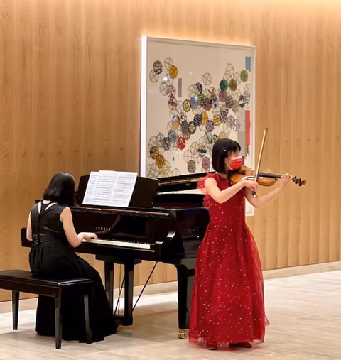 A woman in a red dress and red face mask playing the violin while another woman in a black dress plays a grand piano in a room with wooden paneled walls and abstract artwork hanging behind them.