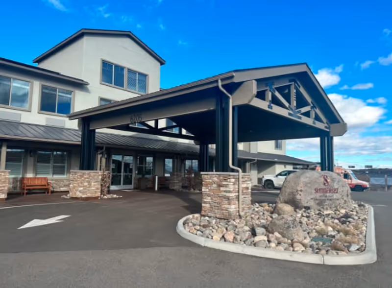 Exterior front entrance of Summerset Senior Living - Memory Care and Adaptive Care facility featuring a covered drop-off area supported by stone and metal pillars, a large rock with the facility's name engraved, a bench near the building, and a clear blue sky with some clouds.