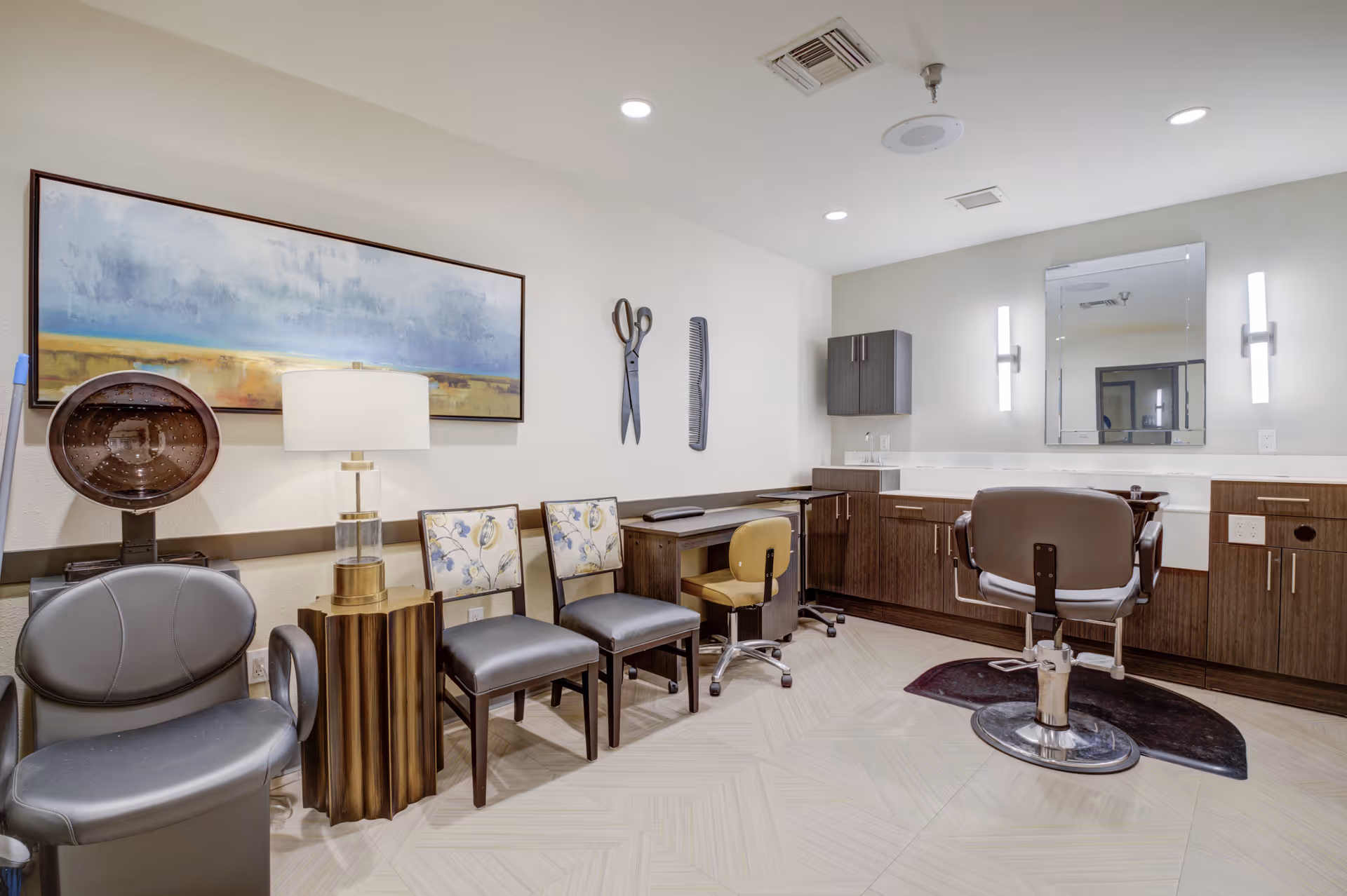 Interior view of a salon or grooming area in an assisted living facility featuring a hair dryer chair, a lamp on a wooden side table, three chairs including two with floral upholstery, a desk with a rolling chair, and a styling chair in front of a large mirror with wall-mounted lights on either side. The walls are decorated with a large landscape painting and oversized wall art of scissors and a comb.