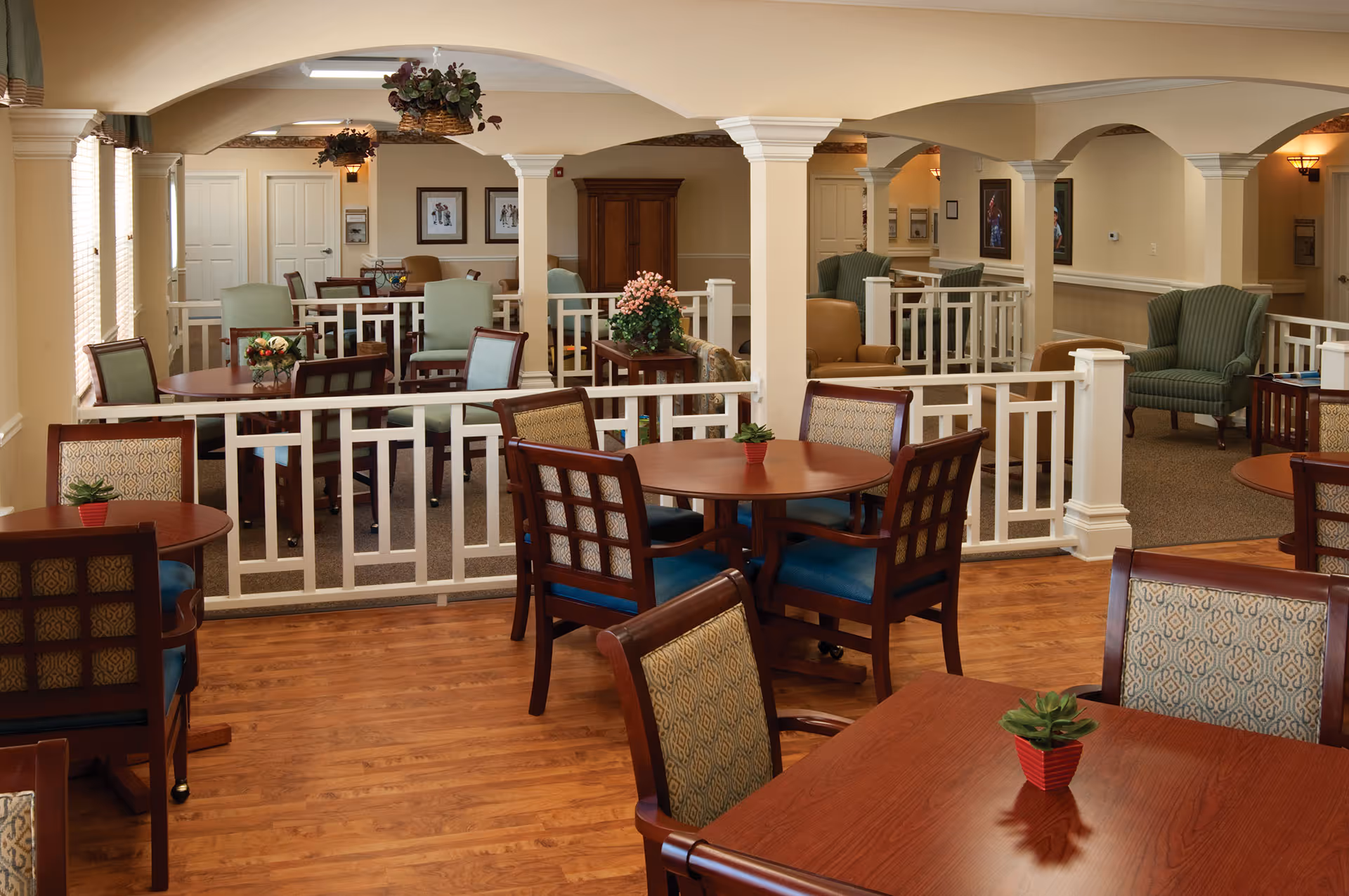 Interior view of a senior living facility common area with multiple round wooden tables and cushioned chairs arranged on a wood floor. The room features white railings, beige walls, arched ceilings, and decorative plants on the tables. Comfortable armchairs and framed artwork are visible in the background.