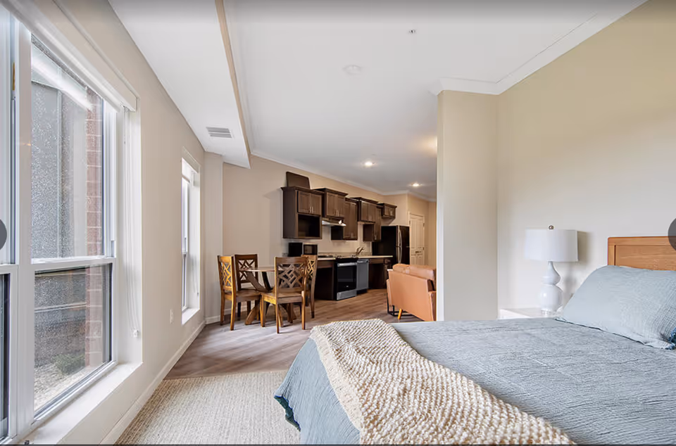 Bright open-plan apartment interior with a bed in the foreground, a bedside table and lamp, a dining table, and a kitchen with dark cabinets.