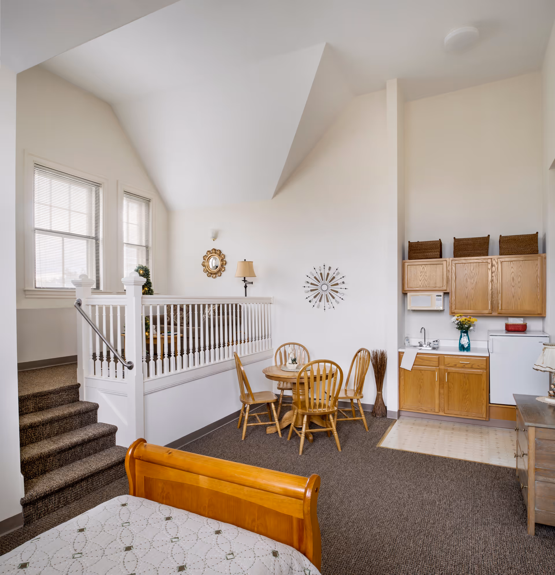 Interior view of a senior living facility room at Corcoran House Assisted Living featuring a wooden bed with patterned bedding in the foreground, a small dining area with a round wooden table and four chairs, a kitchenette with wooden cabinets, a sink, microwave, and mini fridge, and a raised sitting area with a couch and lamp near two windows with blinds.