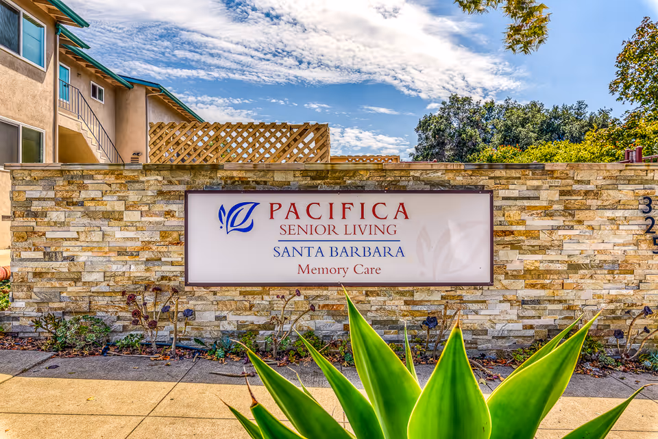 Stone wall with a sign for Pacifica Senior Living Santa Barbara Memory Care, with a building and blue sky in the background and green plants in the foreground.