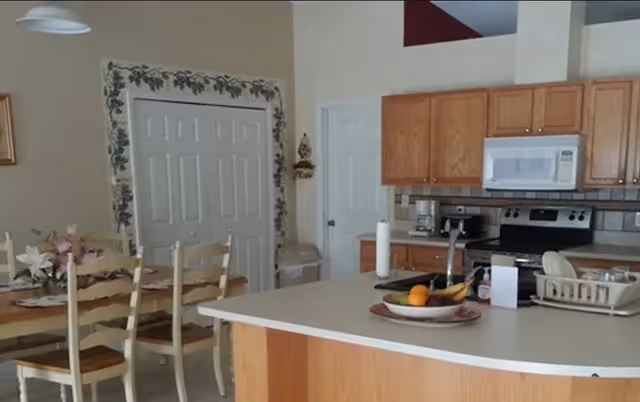 Interior view of a kitchen and dining area with wooden cabinets, a white countertop island with a fruit bowl and dish rack, a stove with a microwave above it, and a dining table with chairs and a floral centerpiece. There are double doors with decorative trim and a trash bin near a closed door.