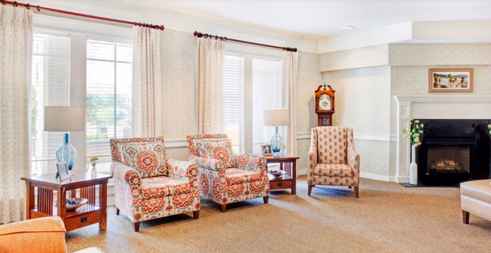Bright living room with patterned armchairs and side tables in front of large windows, a fireplace and a grandfather clock.