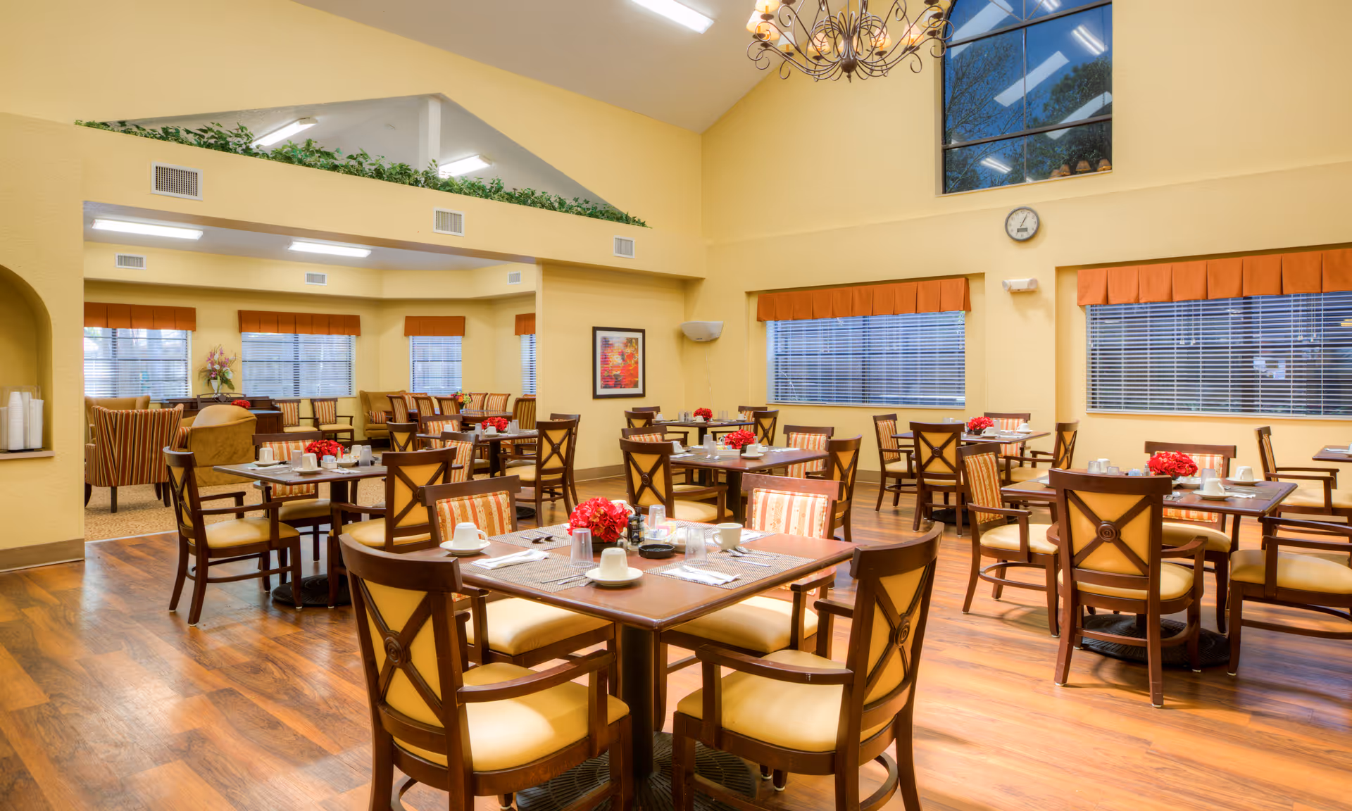 A bright and spacious dining room with multiple wooden tables and chairs arranged neatly. Each table is set with cups, plates, and red floral centerpieces. The room has large windows with orange valances, yellow walls, and a high ceiling with a chandelier. The floor is wooden, and there is a clock on the wall.